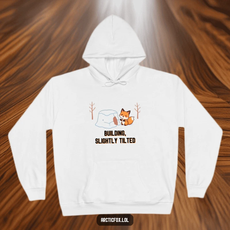 Funny Hoodie: A cheerful fox comically engaged in building a precarious, wobbly snow fort, perfect for winter humor.